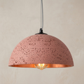 A pendant light unit with a hemispherical design that highlights the raw industrial texture and fine pores, in a warm, earthy pink color that harmonizes with the interior lighting to give a distinctive modern character to the architectural space.